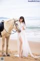 A woman in a white dress standing next to a horse on the beach.